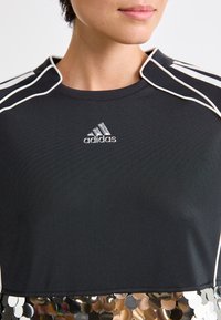 Sieviete valkā melnu Adidas krekli ar baltām svītrām uz pleciem un lielām atstarojošām pērļu detaļām apakšējā malā.