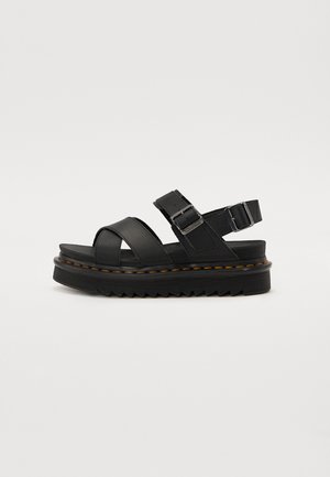 Black platform sandals made of leather, featuring crisscross straps and an adjustable buckled ankle strap. Textured sole with zigzag pattern.