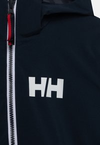 Chaqueta navy con el logo blanco "HH," que presenta un cierre frontal con cremallera y un acento rojo en la cremallera, hecha de tejido suave.