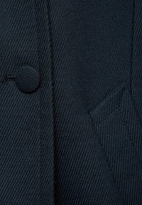 Navy gestructureerde stof Close-up van een ronde knop, zorgvuldig gestikt naad en schuin zakdetail. Het materiaal lijkt duurzaam en geweven.