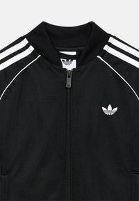 Μαύρο αθλητικό μπουφάν από υφαντό ύφασμα. Διαθέτει λευκές τριπλές ρίγες στους ώμους και μικρό λευκό λογότυπο στο στήθος. Κλείσιμο με φερμουάρ.