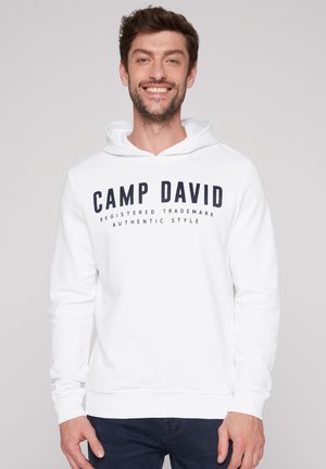 Uśmiechnięty mężczyzna ubrany w białą bluzę z kapturem z napisem "CAMP DAVID", stojący na jednolitym jasnoszarym tle.