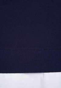 Navy sweater close-up, featuring a ribbed hem and smooth texture, with a contrasting white layer visible underneath.