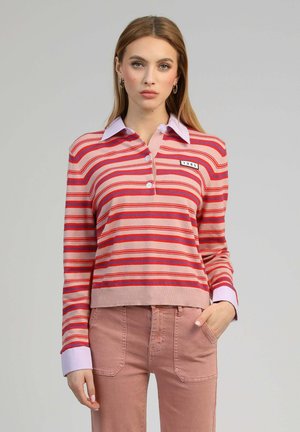 Femme aux cheveux lisses châtain clair portant un pull à col rayé rose, rouge et violet, et un pantalon rose, la main dans la poche.