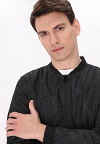 Giubbotto bomber nero con un motivo geometrico, chiusura con zip e polsini a coste. Il materiale appare liscio con una leggera lucentezza. Il modello posa con le braccia incrociate.