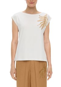Haut blanc à manches courtes avec un décolleté bateau, arborant une broderie en or de fougère sur l'épaule gauche. Associé à un pantalon large beige.