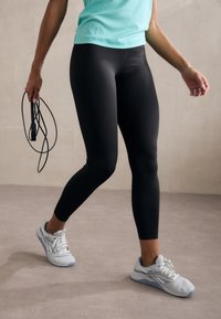 Svarte leggings med en stram passform, kombinert med lyseblå sneakers. Innstillingen har en glatt, nøytral tone på gulvet og veggteksturen.