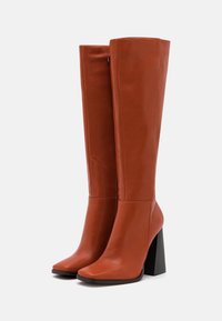 Bottes montantes marron en similicuir avec bout carré, talon noir épais et texture lisse. Design épuré et minimaliste sans ornements.
