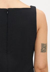 Črna brezroka obleka s kvadratnim izrezom, ki ima zadrgo na hrbtni strani in gladko teksturo. Vidna tetovaža na zgornjem delu roke.