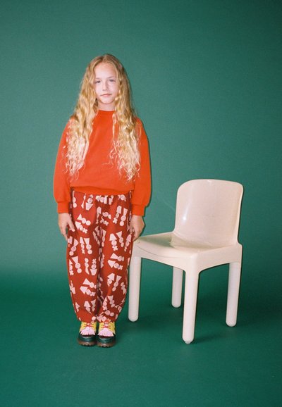 Červená mikina, hnedé nohavice so vzorom geometrických tvarov a farebné topánky. Hned vedľa stojí biela plastová stolička na zelenom pozadí.