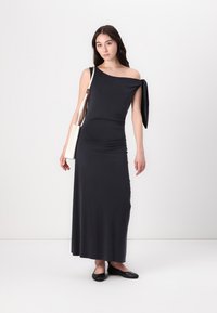 Woman wearing a black one-shoulder long dress with a tie detail, black flats, and carrying a white shoulder bag, standing against a white background.