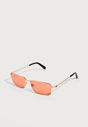 Lunettes de soleil rectangulaires à monture dorée avec des verres teintés pêche et des branches fines à bouts noirs reposant sur une surface claire.