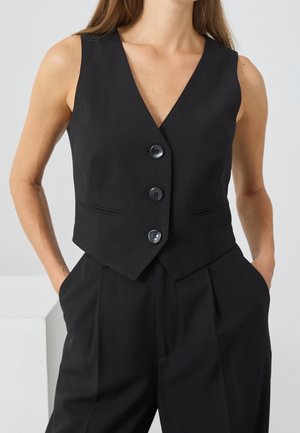 Frau trägt eine eng anliegende schwarze ärmellose Weste mit drei Knöpfen und passende hoch taillierte schwarze Hose mit den Händen in den Taschen.