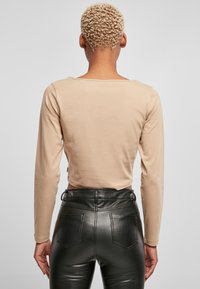 Beiger Langarm-Crop-Top mit weitem Ausschnitt, kombiniert mit hochgeschnittenen schwarzen Kunstlederhosen mit Gesäßtaschen und einer glänzenden Textur.