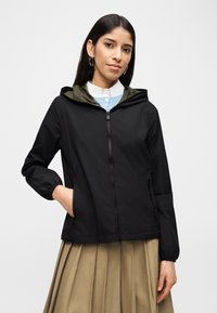 Γυναίκα που φοράει μαύρο φερμουάρ με κουκούλα πάνω από μπλε πουλόβερ και λευκό πουκάμισο με γιακά, με μπεζ πλισέ φούστα και χρυσούς κρίκους σκουλαρίκια.