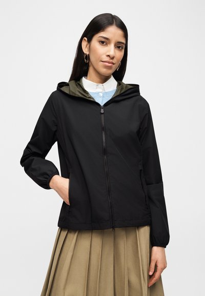 Femme portant une veste à capuche noire zippée par-dessus un pull bleu et une chemise à col blanc, avec une jupe plissée beige et des boucles d'oreilles épaisses dorées.
