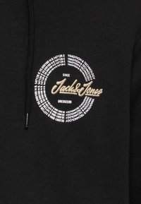 Sudadera negra con un logo circular que presenta "Jack & Jones" en script dorado. El patrón de texto incluye "Tradición y Tecnología" en blanco.