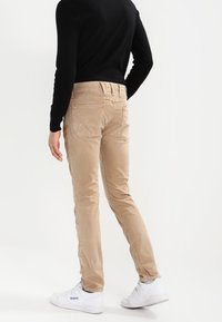 Light beige slim-fit trousers made of cotton blend, featuring back pockets, minimal stitching detail, and paired with white sneakers.