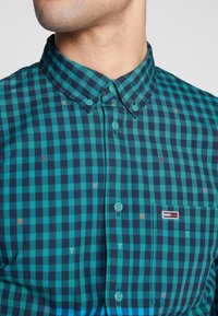 Camicia a maniche corte con bottoni, in motivo a quadri verde petrolio e blu navy, con lettere decorative e una piccola toppa logo sulla tasca del petto.