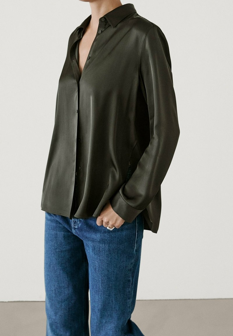 Femme portant une chemise en satin vert foncé boutonnée et un jean bleu, debout une main dans la poche sur un fond uni.