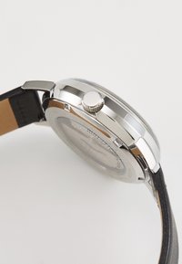 Ceas din metal argintiu cu carcasă rotundă, curea din piele neagră și o coroană texturată; spatele transparent dezvăluie componentele interioare.