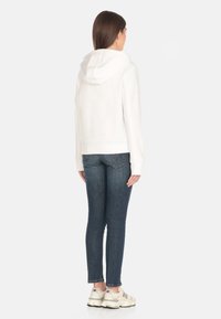 Un sweat à capuche blanc au texture douce, associé à un jean fitted bleu foncé et des baskets blanches. Le design est simple et sans ornement.