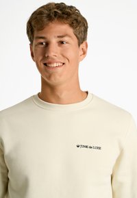 Sudadera color crema hecha de algodón suave, con cuello redondo y un logo bordado en negro que dice "JUNK de LUXE" en el pecho.