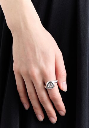 Silberring mit einem kreisförmigen Design und einem klaren Stein, der in der Mitte eingelegt ist, getragen an der Hand einer Person vor einem dunklen Hintergrund.