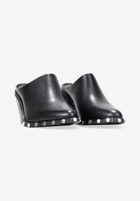 Zuecos de cuero negro con punta redondeada, tacón de cuña y acentos de tachuelas plateadas a lo largo de la suela. Textura suave, diseño minimalista.