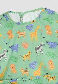 Zelená tkanina so vzorom džungľových zvierat, na ktorej sú levy, žirafy, zebry, tigre a leopardy. Obsahuje aj lístie a geometrické tvary.