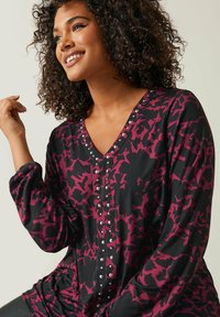 Zwarte blouse met lange mouwen en een abstract maroon patroon, V-hals en decoratieve zilveren studs aan de voorkant. Glad stoffen textuur.