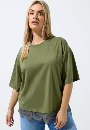 Kvinde med langt blondt hår iført en oversized olivengrøn T-shirt med blonderkant og blå jeans mod en ensfarvet lys baggrund.