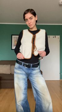 Ivoorkleurige faux fur-vest met bruine biezen en strikjes, gedragen over een zwart shirt, gecombineerd met lichtblauwe, vervaagde jeans met wijde pijpen en een zwarte riem.