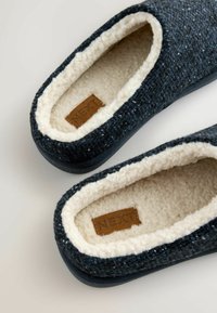 Tmavě modré textilní bačkory s měkkou krémovou podšívkou, robustní černou podrážkou a hnědou etiketou se slovem "NEXT" uvnitř.