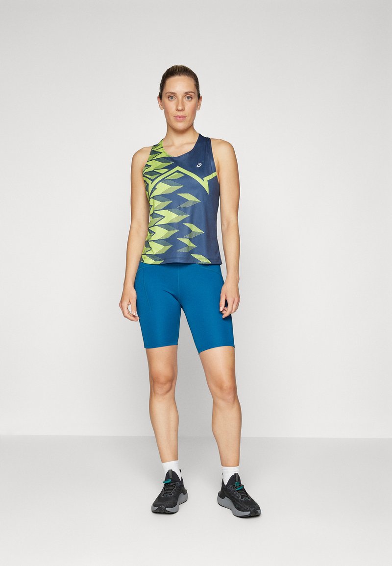 Pantalones cortos deportivos azules con un diseño ajustado, combinados con una camiseta sin mangas que presenta un patrón geométrico verde y marino. Zapatillas deportivas negras.