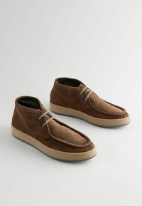 Smeđe antilop cipele do gležnja s načinom moccasin završetka, bež gumeni potplat i sivi vez, s glatkom teksturom i minimalističkim dizajnom.