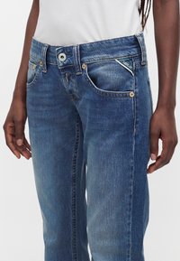 Personne portant un jean Replay bleu avec des poches avant, un bouton visible et une chemise blanche rentrée, debout devant un fond clair uni.