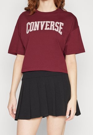 Mujer con una camiseta corta color burdeos con la palabra "CONVERSE" y una minifalda plisada negra contra un fondo liso.