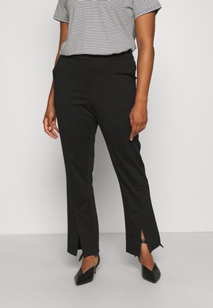 Person iført sorte bukser med lige ben og forreste slidser samt sorte spidse hæle, kombineret med en sort-hvid stribet T-shirt.