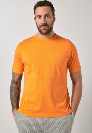 Mężczyzna z brązową brodą i wytatuowaną lewą ręką, ubrany w jaskrawo pomarańską koszulkę z krótkim rękawem oraz pasiastymi spodniami, stoi z rękami w kieszeniach.