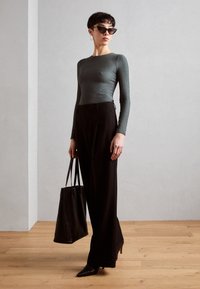 Langärmliges, strukturiertes grünes Oberteil, hochgeschnittene schwarze Hose, schwarze Tote-Tasche, spitze Absatzschuhe und Katzenaugen-Sonnenbrille vor einem minimalistischen Hintergrund.