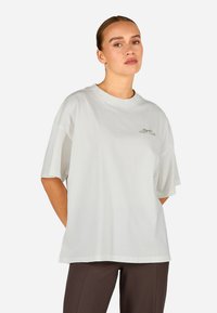 Pārāk liels balts kokvilnas t-krekls ar īsām piedurknēm, apaļo kakla izgriezumu un mazu zeltītu izšūtu "Cozy Club" logotipu uz krūšu.