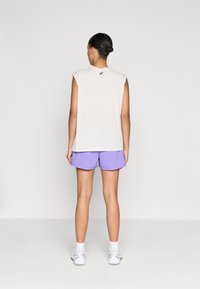 Ανοιχτό γκρι αμάνικο τοπ με λογότυπο στην πλάτη, συνδυασμένο με λιλά σορτς και λευκά αθλητικά παπούτσια, σε απλό φόντο.