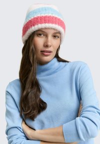 Bonnet en tricot avec des rayures rose, bleu et blanc. Texture douce. Modèle portant un col roulé bleu clair, cheveux mi-longs. Fond neutre.