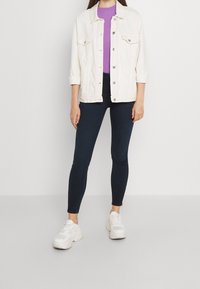 Chaqueta de mezclilla clara, top de canalé púrpura, leggings oscuros y zapatillas blancas. La chaqueta tiene botones plateados y dos bolsillos en el pecho.