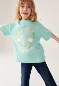 Mintgrøn t-shirt med korte ærmer, der har et farverigt smilende ansigt design, regnbue og teksten "GLAD & VENLIG siden 1974." Sammen med mørke jeans.