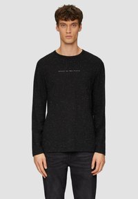 Schwarzes Langarmshirt mit einem gesprenkelten Muster, Rundhalsausschnitt und dezentem Textdetail auf der Vorderseite. Kombiniert mit dunklen Jeans.