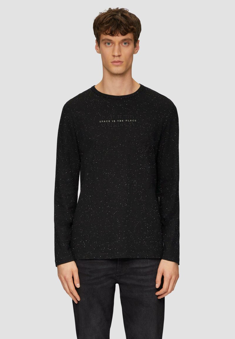 Schwarzes Langarmshirt mit einem gesprenkelten Muster, Rundhalsausschnitt und dezentem Textdetail auf der Vorderseite. Kombiniert mit dunklen Jeans.