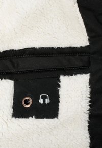 Étiquette en tissu noir avec une icône blanche de casque audio et un œillet métallique attaché à un tissu blanc duveteux et texturé.