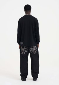 Schwarzes Langarmshirt mit lockerer Passform, kombiniert mit schwarzen Hosen, die über weiße Spinnennetzstickerei auf den hinteren Taschen verfügen.